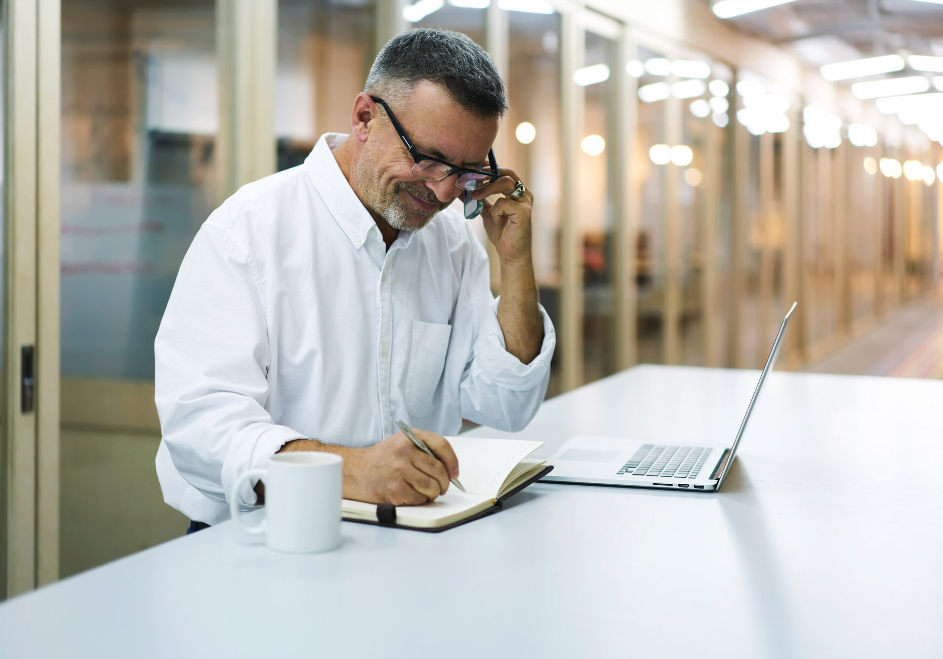 Handsome mature businessman in eyewear noting information from web page browsed on smartphone, serious executive manager concentrated on writing contact from mobile in notebook standing in office Handsome mature businessman in eyewear noting information from web page browsed on smartphone, serious executive manager concentrated on writing contact from mobile in notebook standing in office